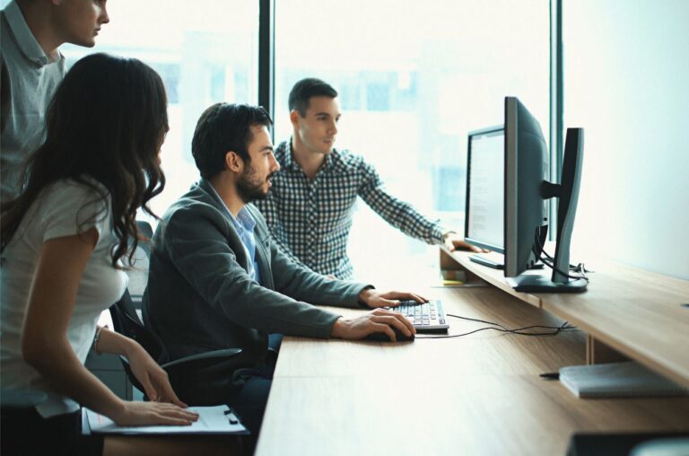 A group of business professionals gather around a two wiedscreen monitors.